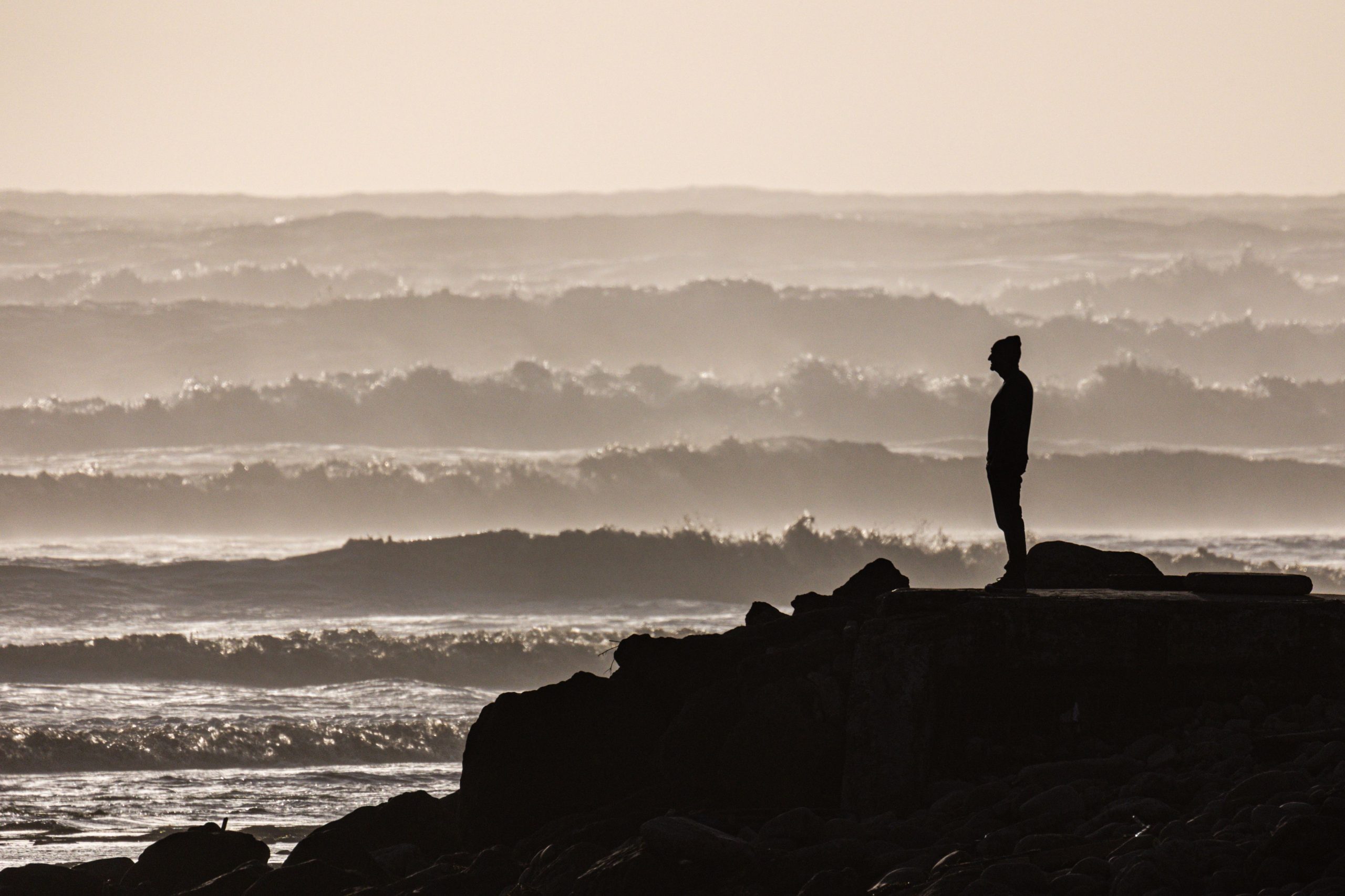 a silhouetted lone figure on a rocky outcrop looking out over a monochrome sea.